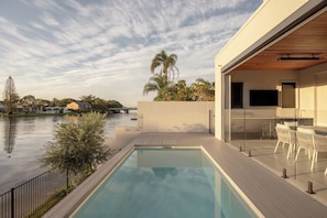 A modern outdoor pool area with a view of a river, a wooden deck, and a glass-enclosed dining area. - Maison by Khove (Broadbeach Waters)