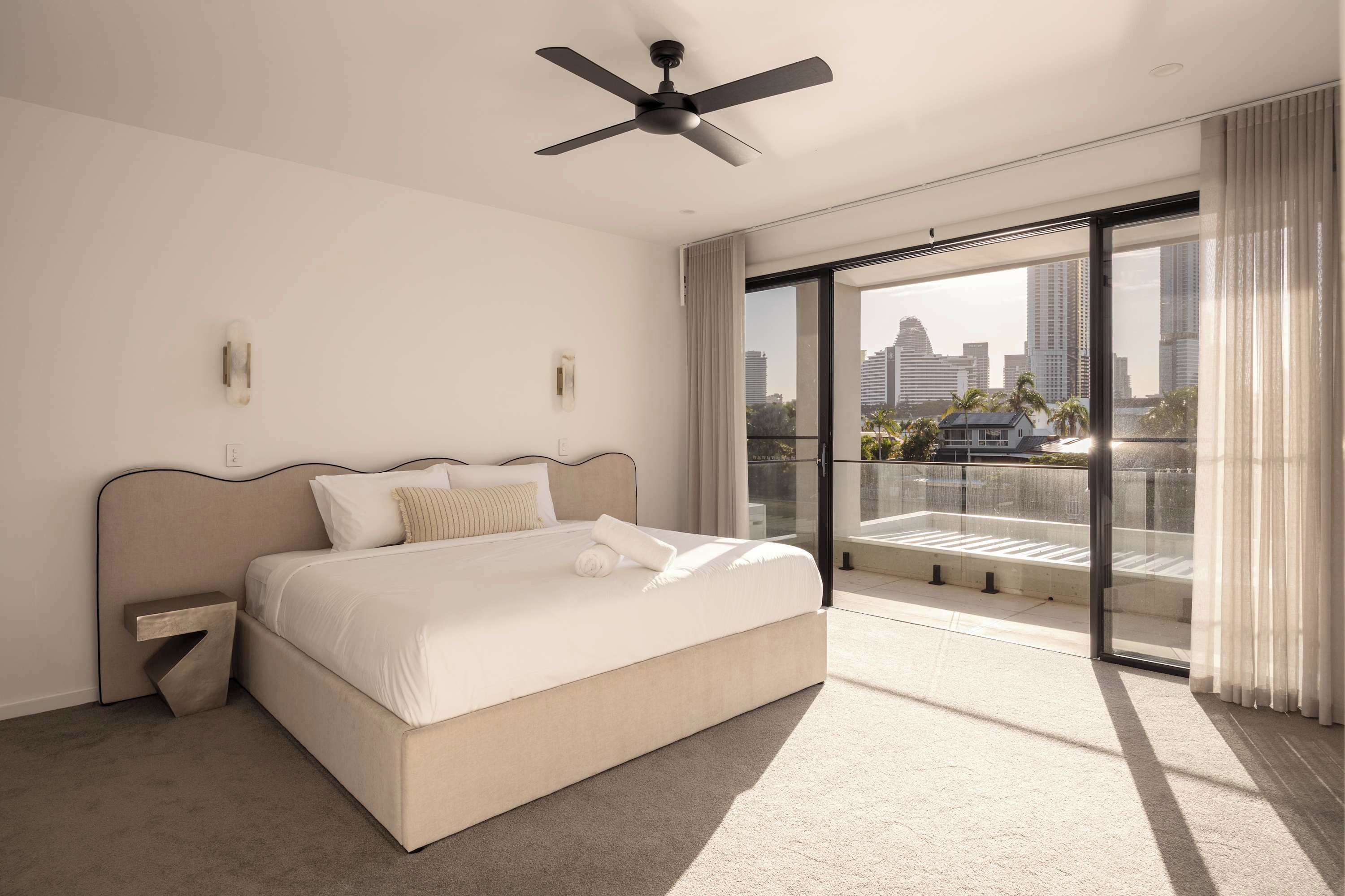 A modern hotel room with a bed, a ceiling fan, a large window with a city view, and a balcony.