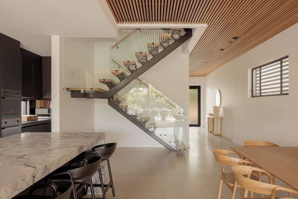 A modern kitchen with a marble countertop and bar stools, a staircase with glass railings, and a dining area with wooden chairs.
