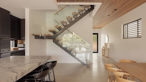 Una cocina moderna con encimera de mármol y taburetes de bar, una escalera con barandilla de cristal y un comedor con sillas de madera.