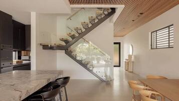 A modern kitchen with a marble countertop and bar stools, a staircase with glass railings, and a dining area with wooden chairs.