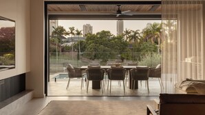 A modern room with a large sliding glass door leading to an outdoor dining area with a pool, surrounded by greenery and cityscape views. - Maison by Khove (Broadbeach Waters)