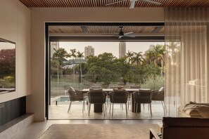 Una habitación moderna con un amplio ventanal corredero que da a un área de comedor exterior con piscina, rodeada de vegetación y vistas de la ciudad.