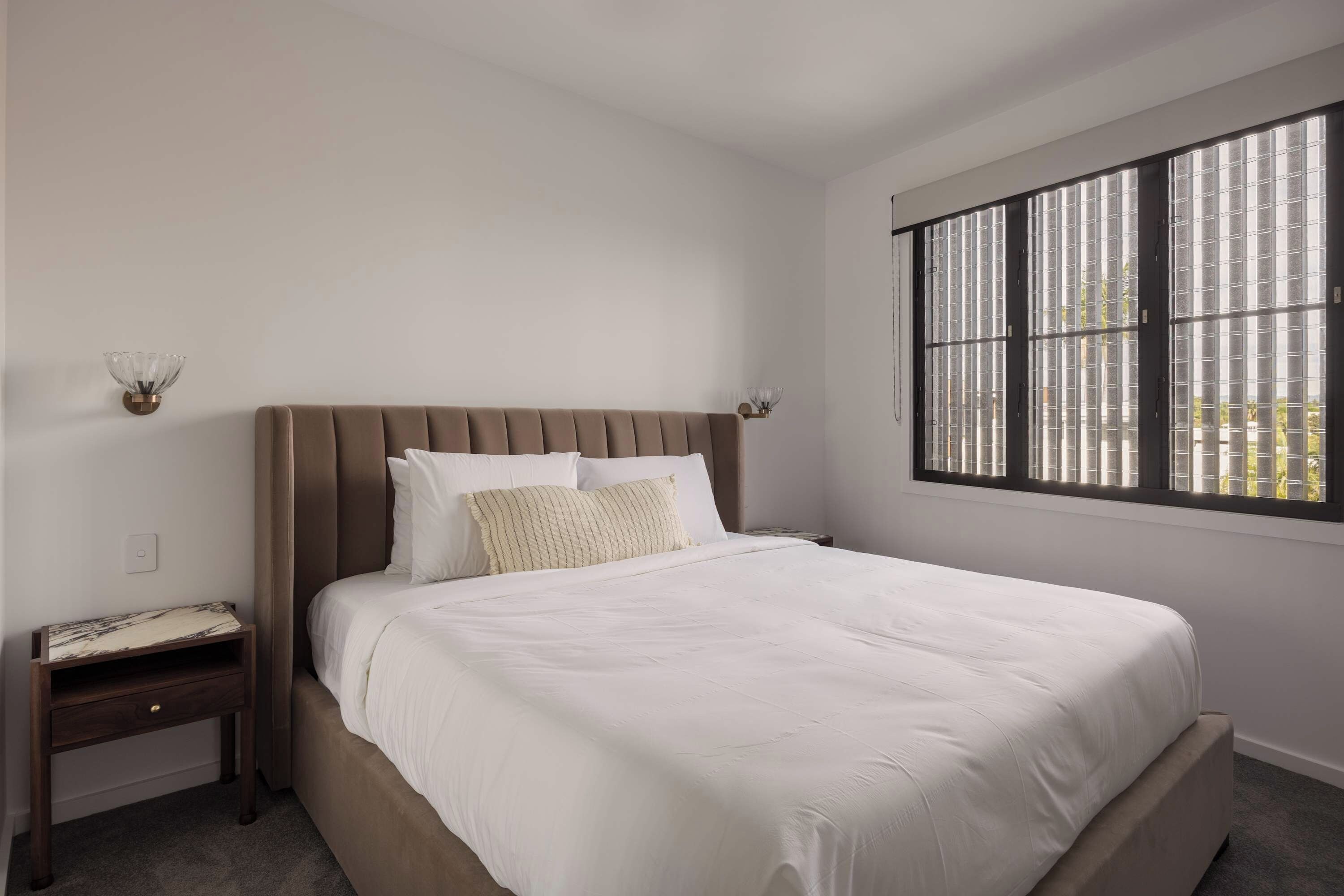 A bedroom with a large bed, a nightstand, a wall-mounted lamp, and a window with blinds.