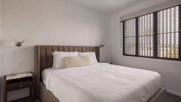 A bedroom with a large bed, a nightstand, a wall-mounted lamp, and a window with blinds.
