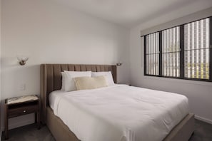 A bedroom with a large bed, a nightstand, a wall-mounted lamp, and a window with blinds. - Maison by Khove (Broadbeach Waters)