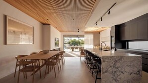 A modern kitchen with a marble island, wooden ceiling, and a dining area with chairs and a table. - Maison by Khove (Broadbeach Waters)