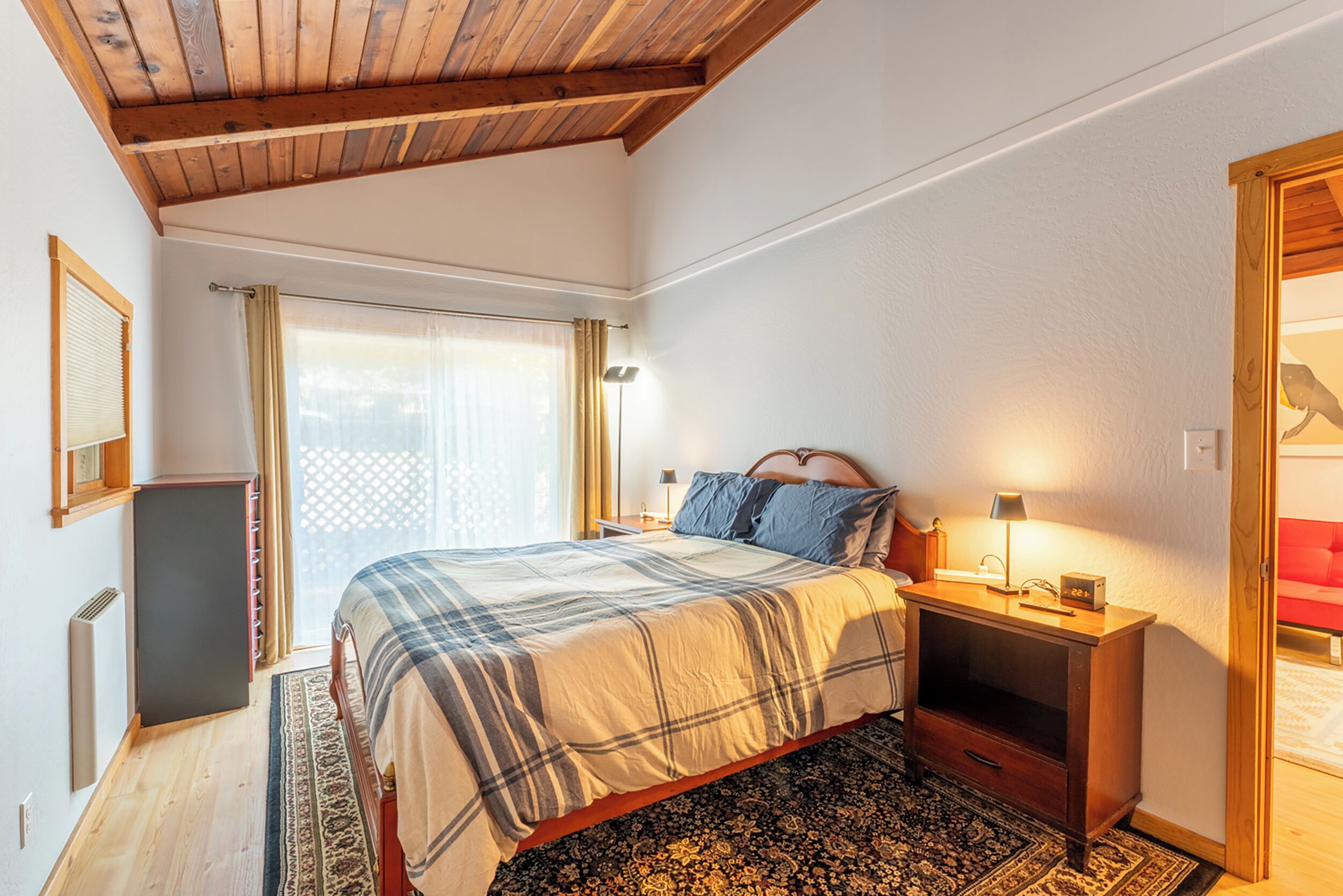 A bedroom with a bed, bedside tables, a nightstand, a lamp, a window with curtains, and a wooden ceiling.