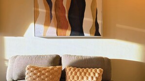 A room with a wall-mounted abstract painting, a sofa with two cushions, and a window allowing natural light.