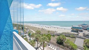 A beach view from a balcony with a clear sky, a wooden boardwalk, and a beach area with umbrellas and people. - Amazing Large Private Studio Condo with Amazing View (Myrtle Beach)