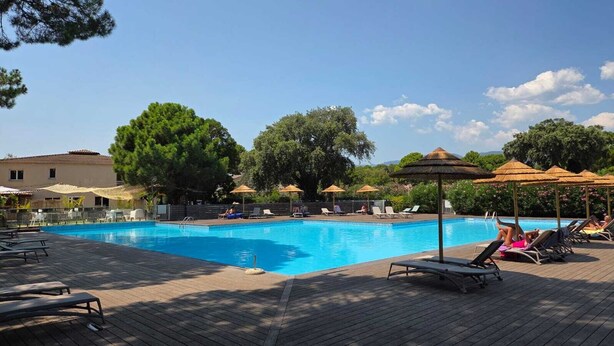 Piscina com espreguiçadeiras e guarda-sóis, cercada por árvores e um edifício.