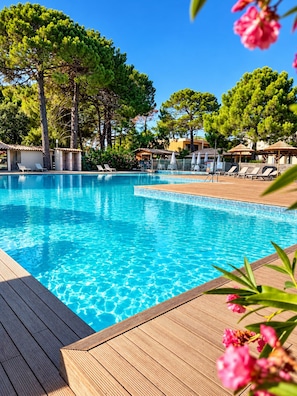 Une piscine d’un bleu éclatant, dotée d’une terrasse en bois et de chaises longues, entourée d’arbres et bordée d’un bâtiment au toit recouvert de tuiles.