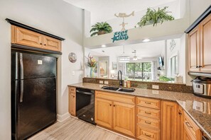 A kitchen with wooden cabinets, a black refrigerator, a dishwasher, a granite countertop, and a hanging plant. - New 2BR, Lake View with Amazing Pool (Myrtle Beach)