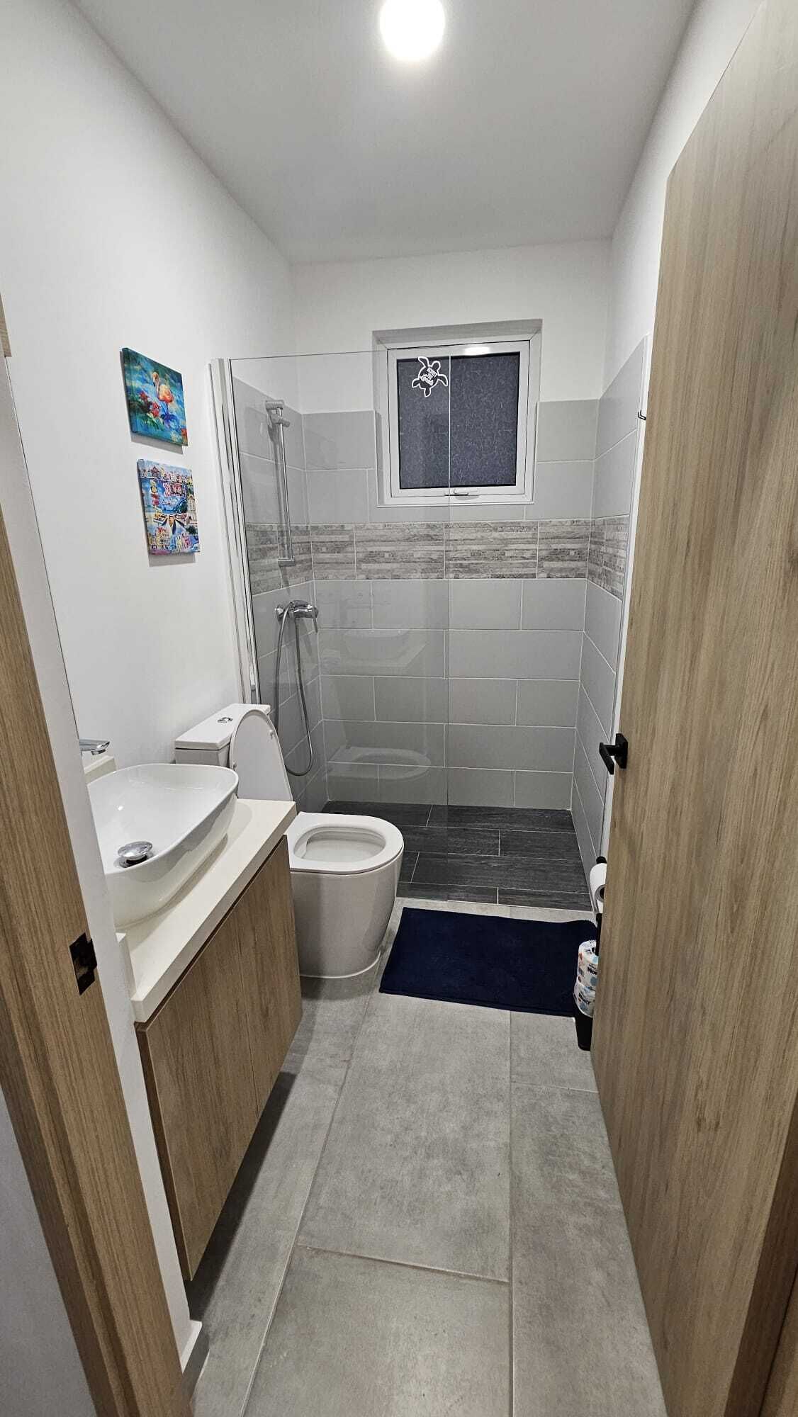 Un baño con inodoro, bidé y lavamanos, ducha con puerta de vidrio y una pequeña ventana sobre la ducha.