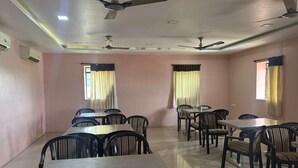 Una sala de conferències amb diverses taules i cadires, ventiladors al sostre i finestres amb cortines.