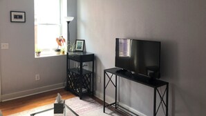 Un salon avec un téléviseur posé sur un meuble, une petite table avec une lampe et une fenêtre ornée de plantes.