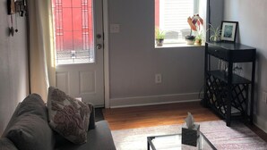 Un salon avec un canapé gris, une table basse en verre, un ventilateur de plafond, une fenêtre donnant sur un beau paysage et une porte avec un portail rouge.