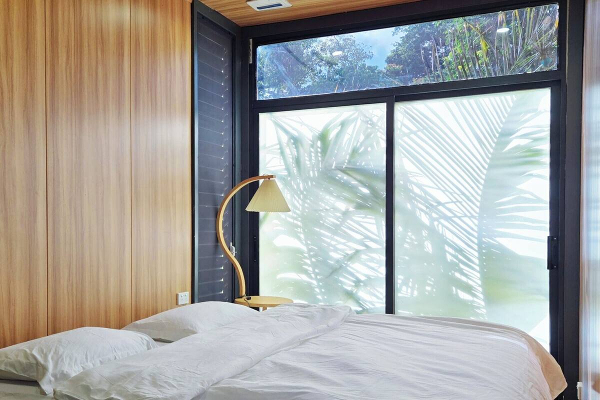 A bedroom with a wooden ceiling, a bed with white bedding, a floor lamp, and a large window with a view of trees.