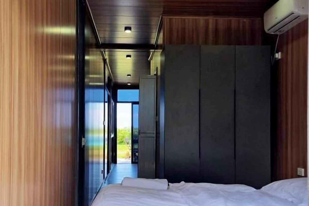 A bedroom with a bed, wooden walls, a dark wardrobe, and a window with a view.