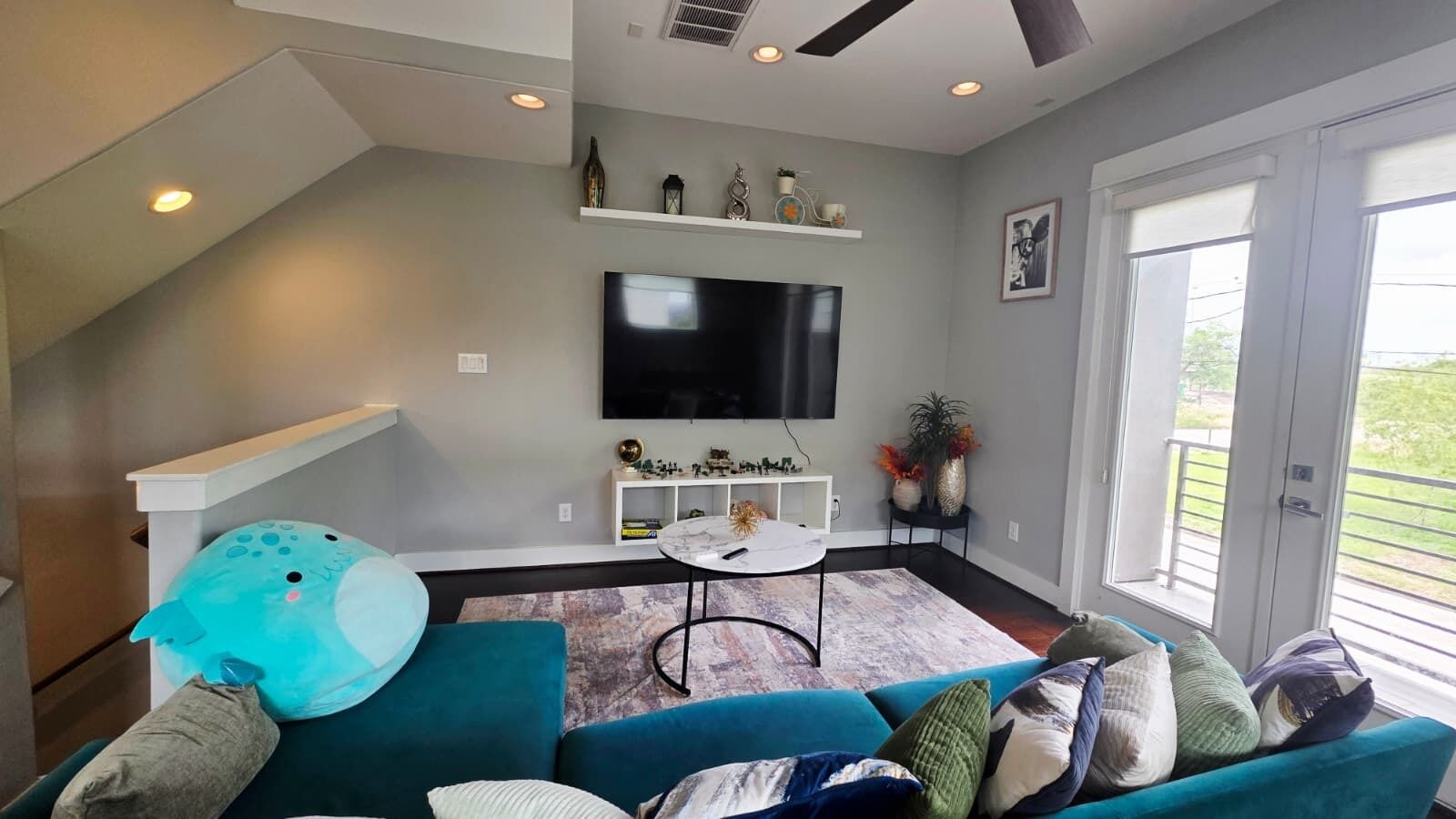 Ein modernes Wohnzimmer mit einem türkisblauen Sofa, einem Couchtisch, einem flachbildfernseher an der Wand und einer kleinen weißen Regal mit Dekorationselementen.
