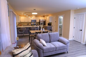 A modern living room with a grey sofa, a dining area with a table and chairs, a kitchen with white cabinets and appliances, and a door leading to another room. - Wonderful Entire home in Kent 3-bedroom with AC in cool walk in train station (Kent)