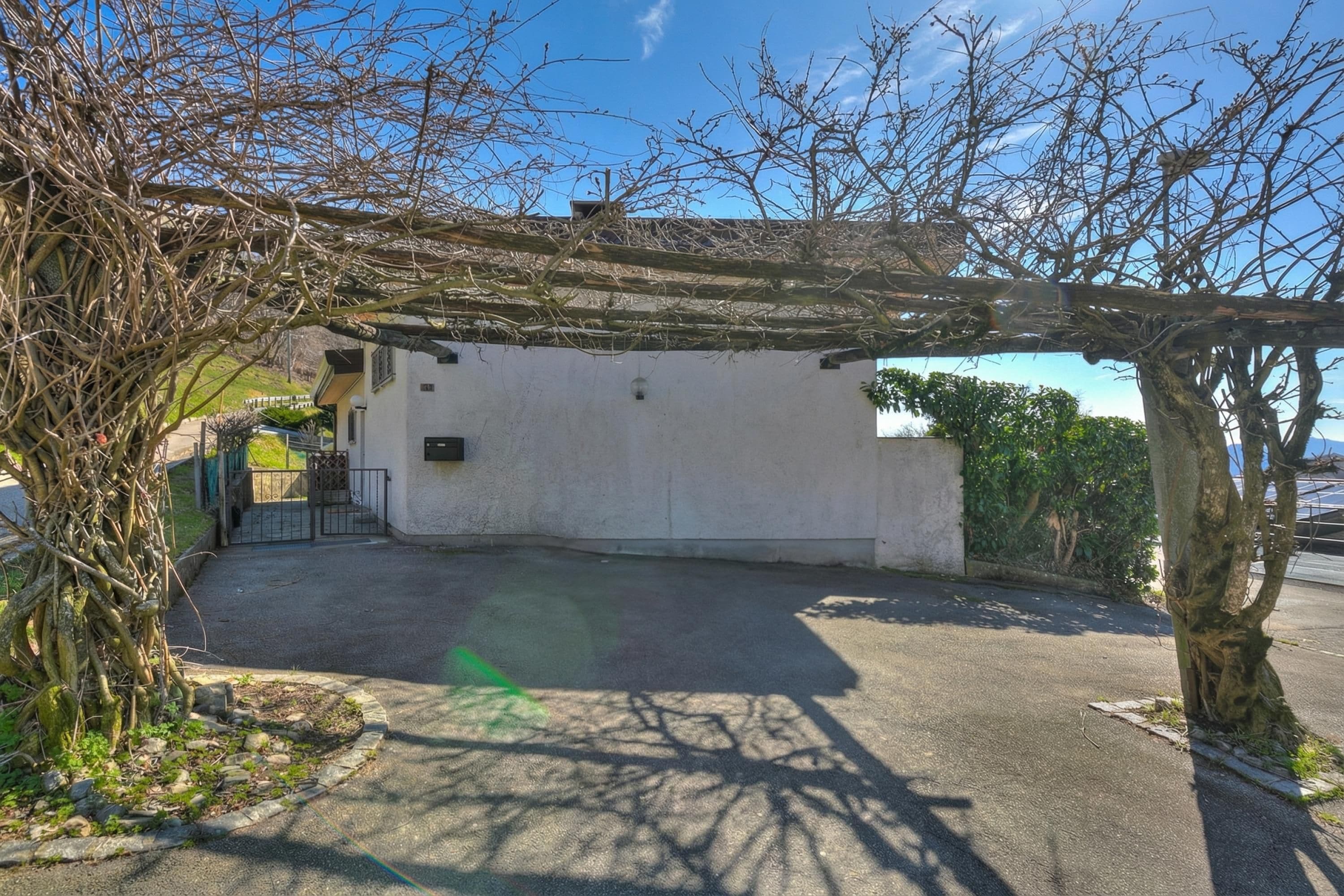 A covered outdoor area with a concrete floor, a pergola, and a white building with a door.