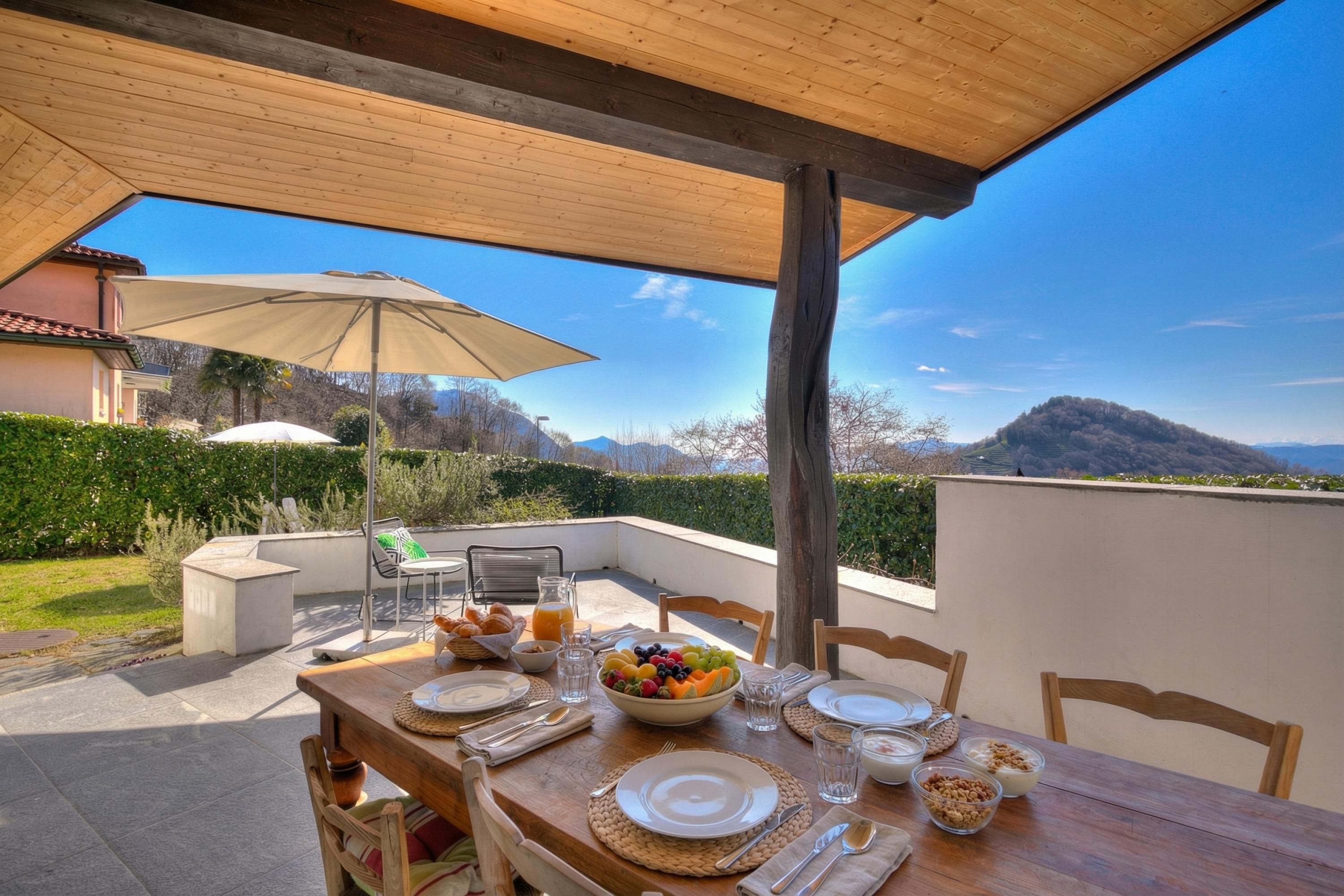 An outdoor dining area with a table set for breakfast, a wooden pergola overhead, and a scenic mountain view in the background.