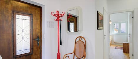 Una puerta de madera con un panel de vidrio decorativo, un perchero rojo, una silla de mimbre, un espejo y un pasillo que conduce a otra habitación.
