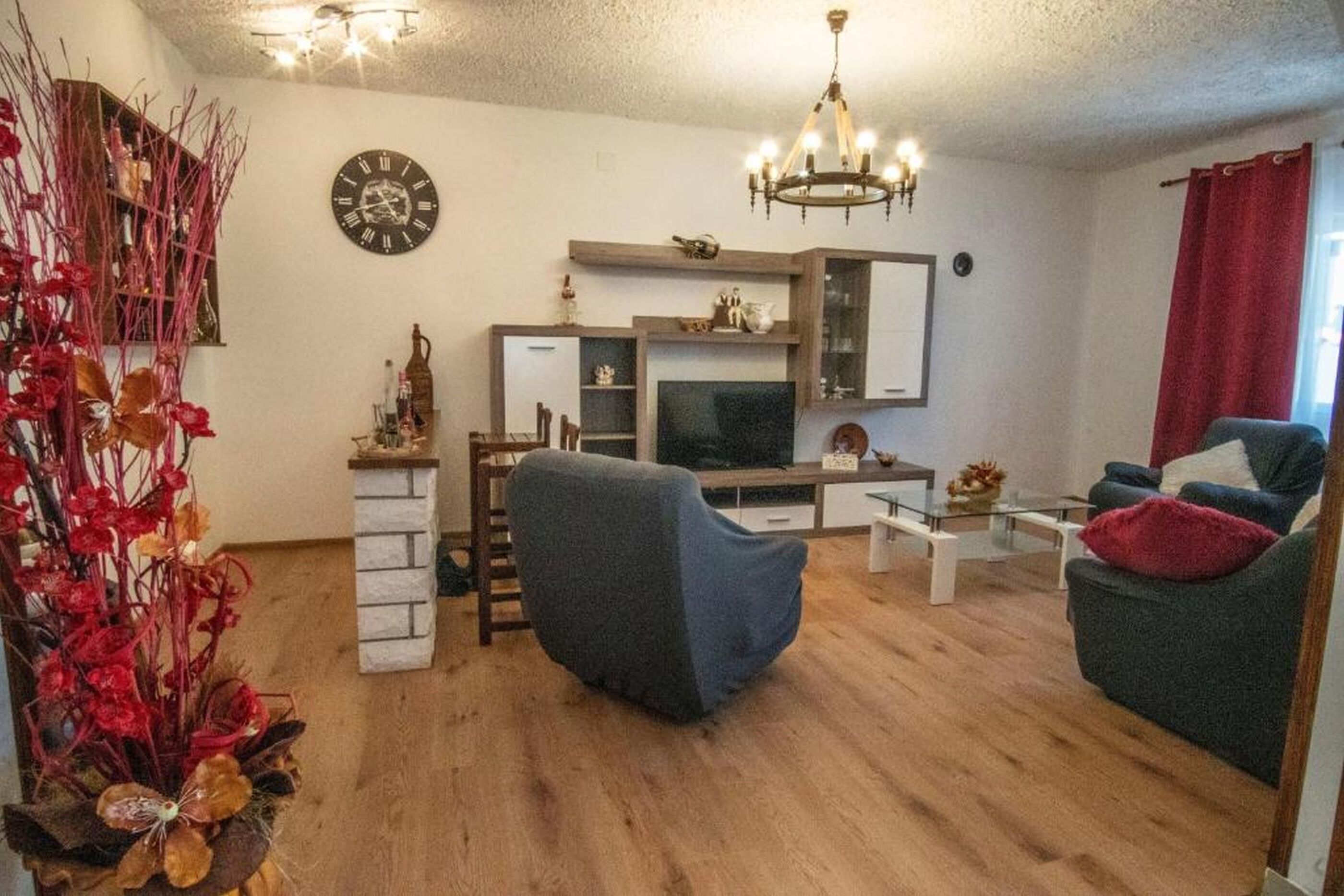 A living room with a TV, a chandelier, a clock, and a red curtain.