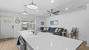 Eine moderne Küche mit einer zentralen Kochinsel, Pendelleuchten, einem Deckenventilator und Blick ins Freie durch große Fenster.