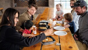 Un groupe de personnes est assis autour d’une table en bois, devant des plats et un gaufre.