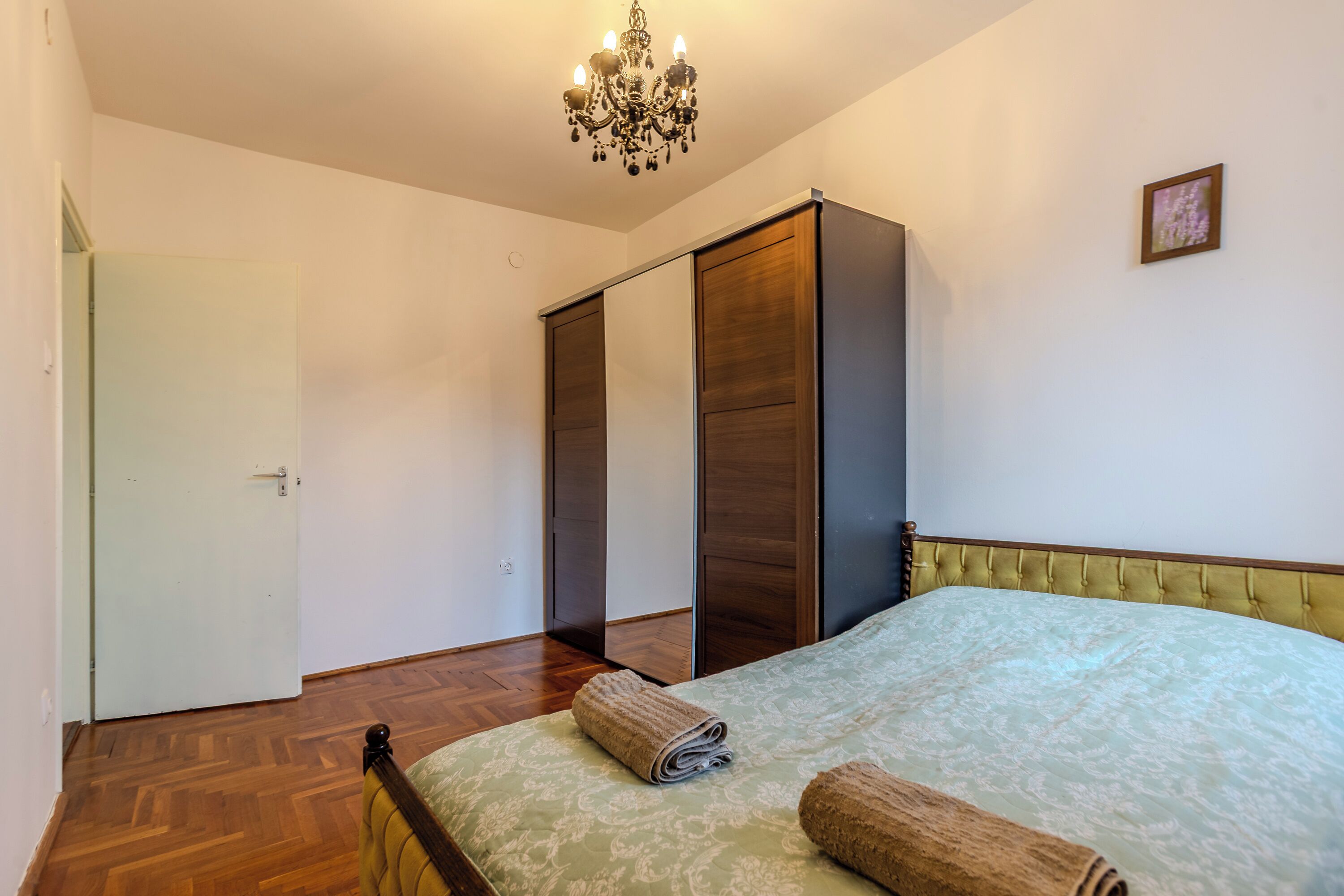 A bedroom with a bed, a wooden wardrobe, a chandelier, and a door.