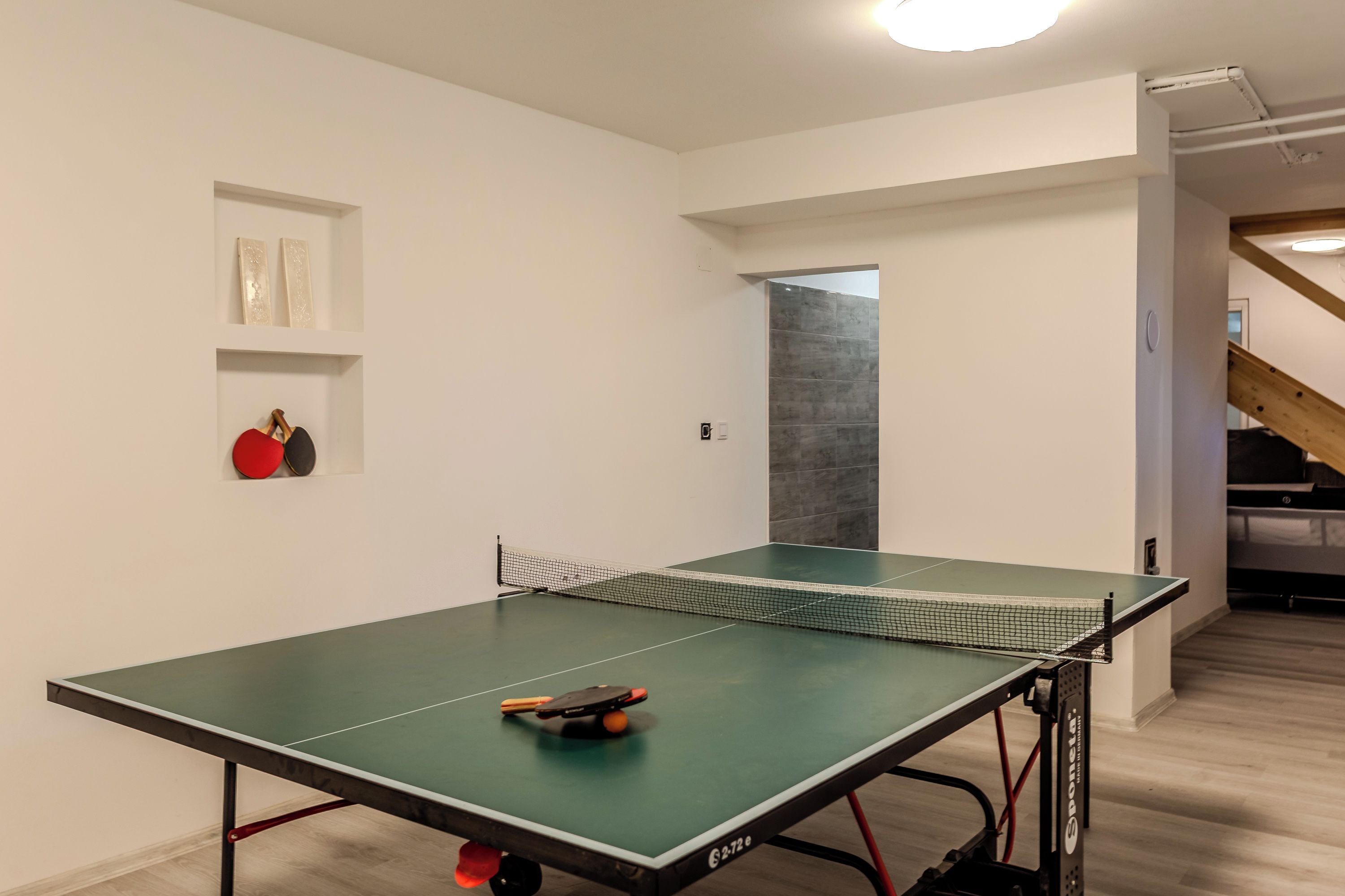 Uma mesa de tênis de mesa em um quarto com paredes brancas, piso de madeira e uma escada ao fundo.