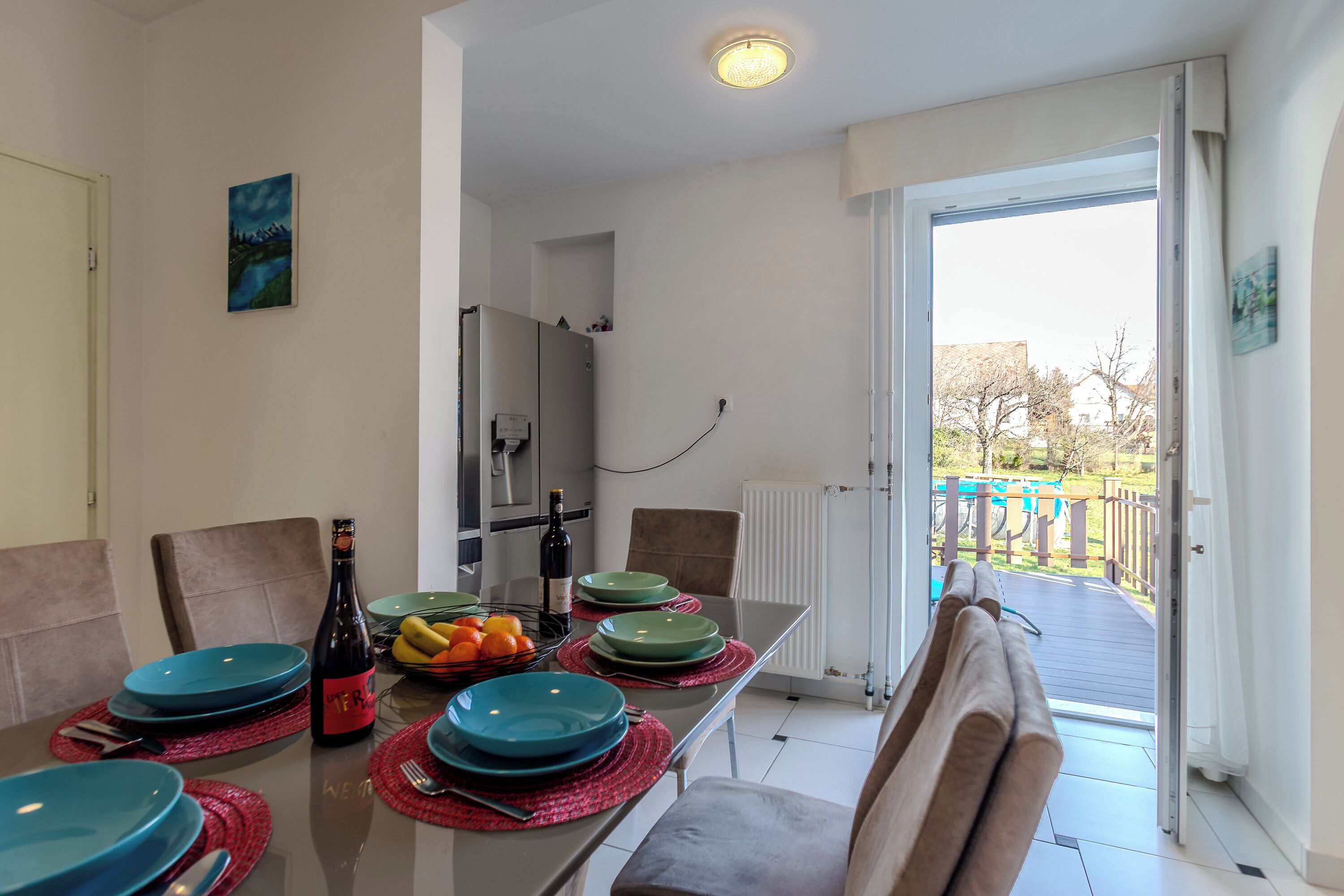 A dining area with a table set for four, a refrigerator, and a view of the outside.