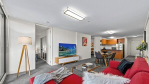 A modern living room with a red sofa, a TV, and a dining area with a kitchen in the background. - Exclusive Stays - The Summit (Southbank)