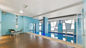 A modern indoor swimming pool with clear glass walls, wooden flooring, and a seating area with chairs. - Exclusive Stays - The Summit (Southbank)