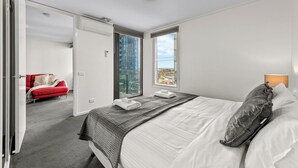 A bedroom with a large bed, a red chair, a window with a city view, and a door leading to another room. - Exclusive Stays - The Summit (Southbank)