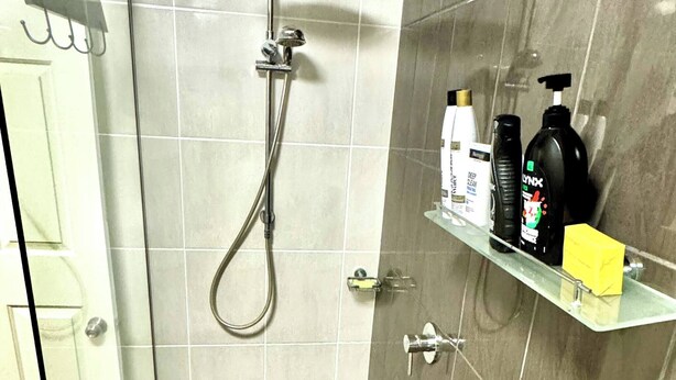 A shower with a handheld showerhead, a glass door, and a shelf with toiletries.