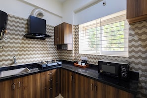 A kitchen with wooden cabinets, a black countertop, a microwave, and a window with blinds. - SLV Ventures Anagha by Alaya Stays 1-BHK (Bengaluru)