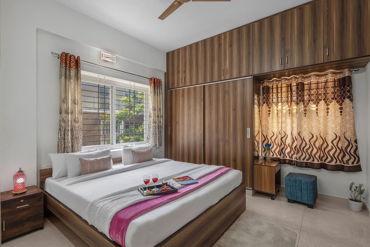 Un dormitorio con cama, armario de madera, ventana con cortinas, mesita de noche y tocador.