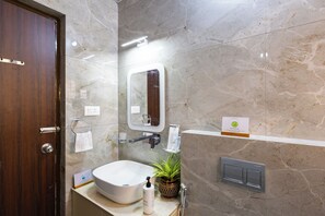 A bathroom with a marble wall, a wooden door, a sink, a mirror, a towel rack, a shelf with toiletries, and a thermostat. - SLV Ventures Anagha by Alaya Stays 1-BHK (Bengaluru)