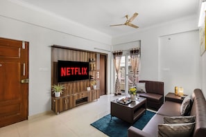 A modern living room with a flat-screen TV, a ceiling fan, and a large window with curtains. - SLV Ventures Anagha by Alaya Stays 1-BHK (Bengaluru)
