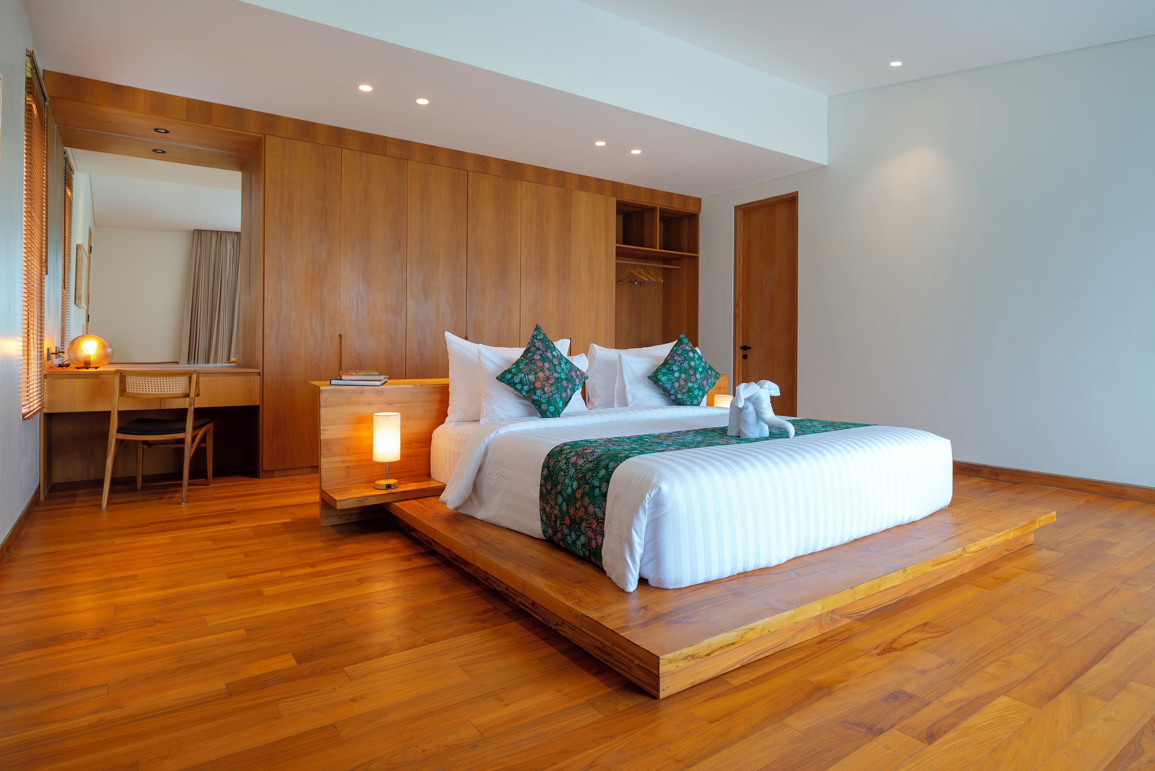 A modern bedroom with a wooden floor, a bed with white bedding and green pillows, a desk with a chair, and a large mirror.