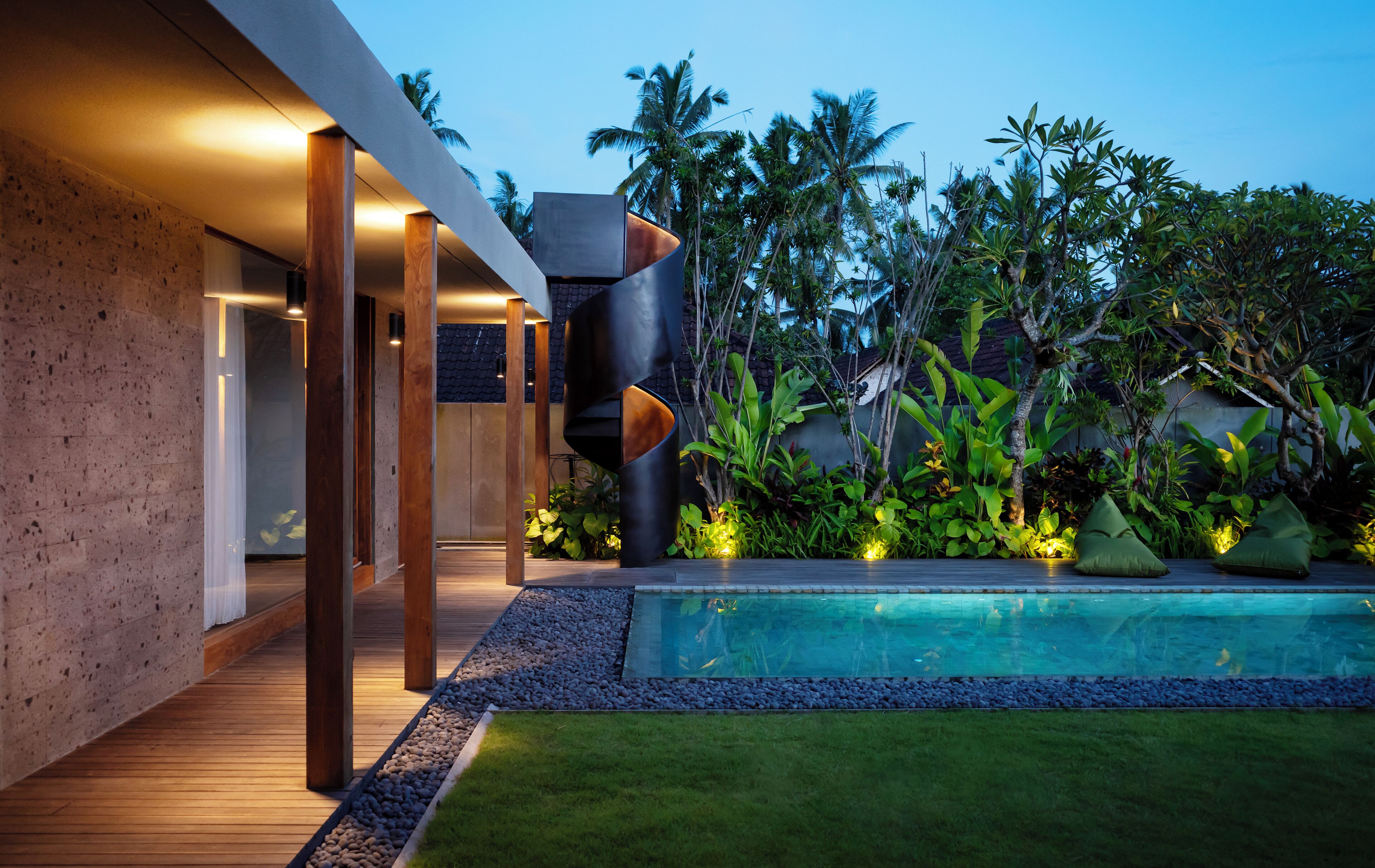A modern outdoor pool area with a wooden deck, a curved black structure, and tropical vegetation.