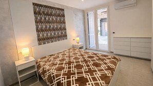 A bedroom with a bed, bedside tables, a wall-mounted artwork, a sliding glass door to the outside, and a built-in air conditioning unit. - Cristallo Bilo 1 al Cavo (Rio Marina)