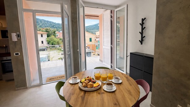 A dining area with a wooden table, breakfast items, and a view of a residential area through open doors. - Cristallo Bilo 1 al Cavo (Rio Marina)