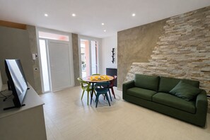 Ein modernes Wohnzimmer mit einem grünen Sofa, einem Esstisch mit bunten Stühlen, einem Flachbildfernseher und einer Wand mit Steinoptik.