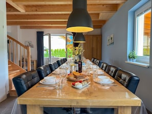 Un espace repas avec une longue table en bois, des chaises en cuir noir, un lustre et une vue sur la verdure à l’extérieur.