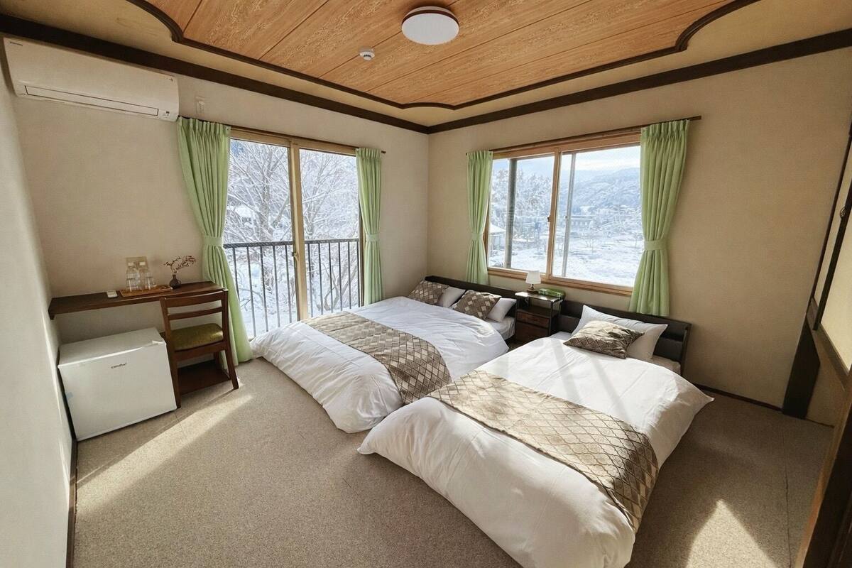 A hotel room with two beds, a desk, a small refrigerator, and large windows with curtains.