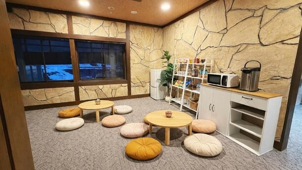 A cozy room with a stone wall, a window with a view, a kitchenette, and several round tables with cushions. - START HOUSE PIAA-Ski IN Ski OUT (Minamiuonuma)